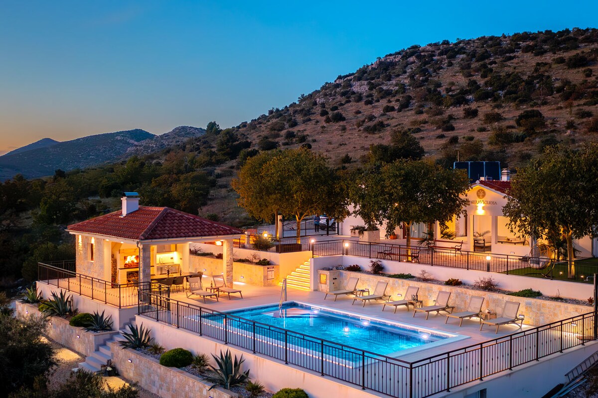 A spacious villa is set against a backdrop of hills, with an outdoor pool illuminated at dusk. Surrounding the pool, lounge chairs are positioned for relaxation, while a covered seating area features a stone structure. Lush greenery enhances the tranquil atmosphere.