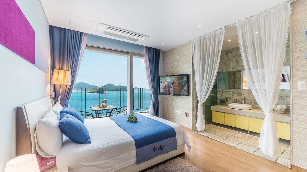 A bright and airy room features a spacious bed with blue accents. Large windows showcase views of the ocean and distant hills. A stylish bathroom area is visible, separated by sheer curtains, with modern fixtures and a relaxing bathtub.