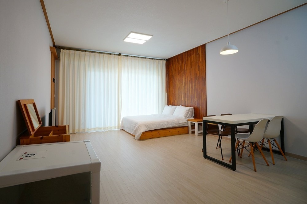 A spacious studio features a double bed against a wooden accent wall. Large windows draped with sheer curtains allow natural light to fill the room. A dining table with four chairs is positioned nearby, complemented by a small storage unit.