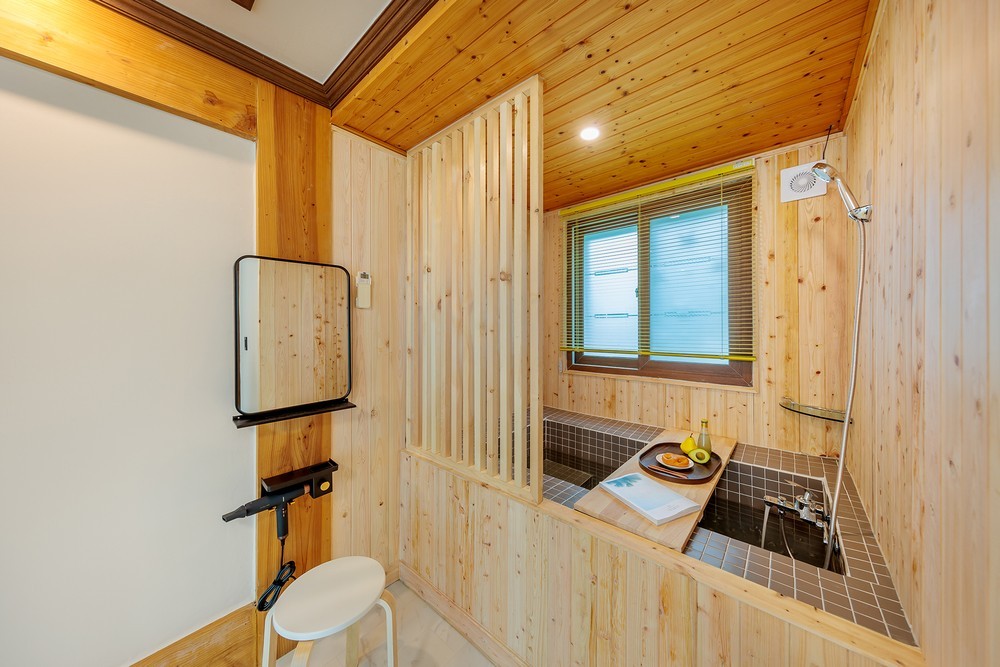 A wooden-paneled bathroom features a private soaking tub designed for relaxation. Natural light filters through two windows, enhancing the serene ambiance. A small table is positioned nearby with a plate of fruit, and a mirror is mounted on the wall.