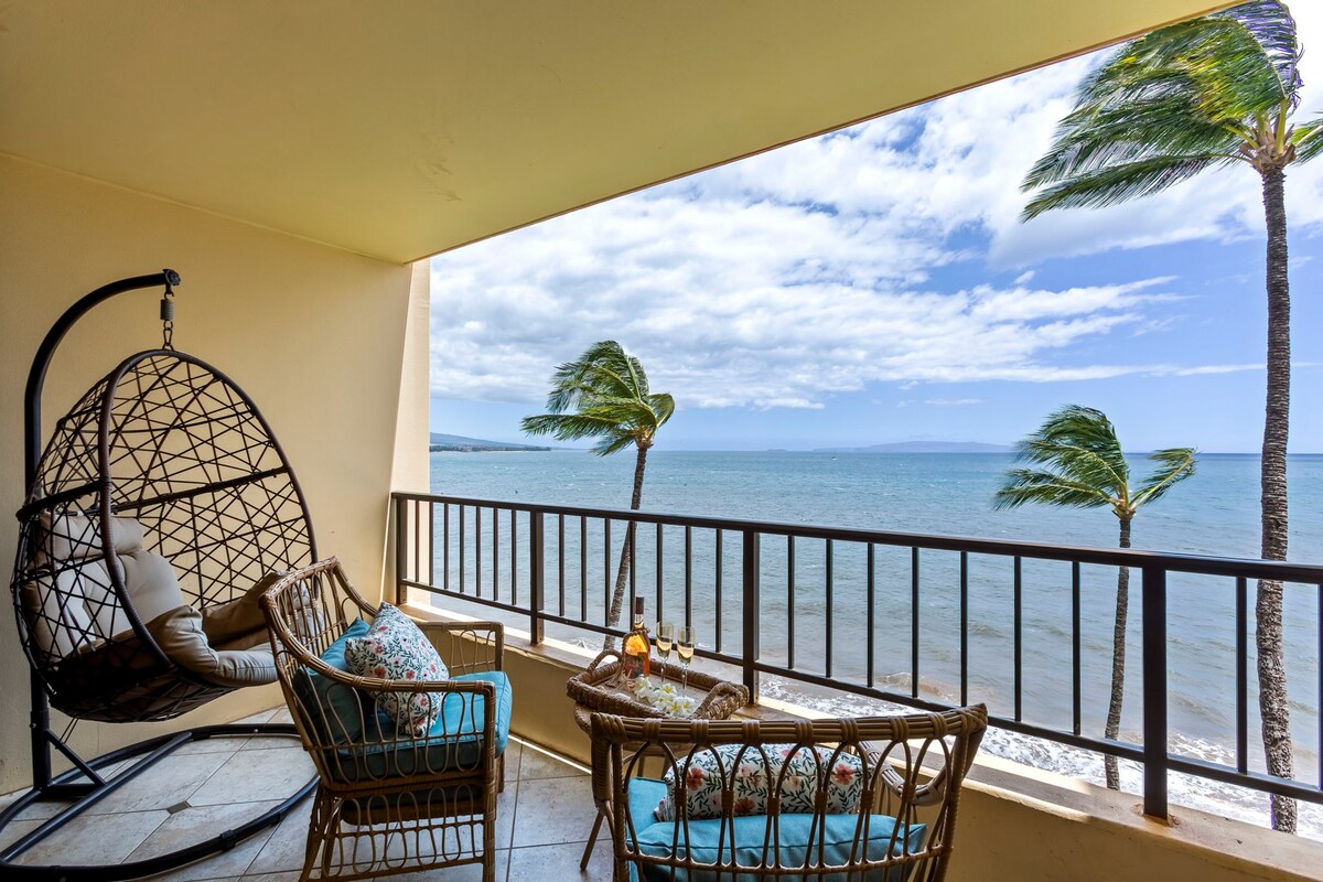 A private lanai is showcased with ocean views, featuring a hanging egg swing and two rattan chairs. The scene includes swaying palm trees against a backdrop of open water and a partly cloudy sky, offering a serene outdoor space for relaxation.