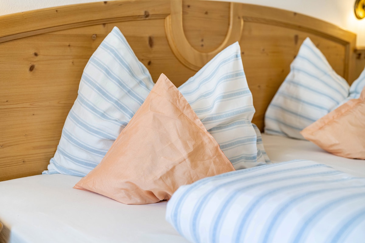 A light and airy bedding arrangement features a wooden headboard with multiple cushions. The striped blue and white pillows are complemented by a peach-colored accent pillow. The spacious bed is dressed in crisp white linens, allowing a serene and comfortable atmosphere.