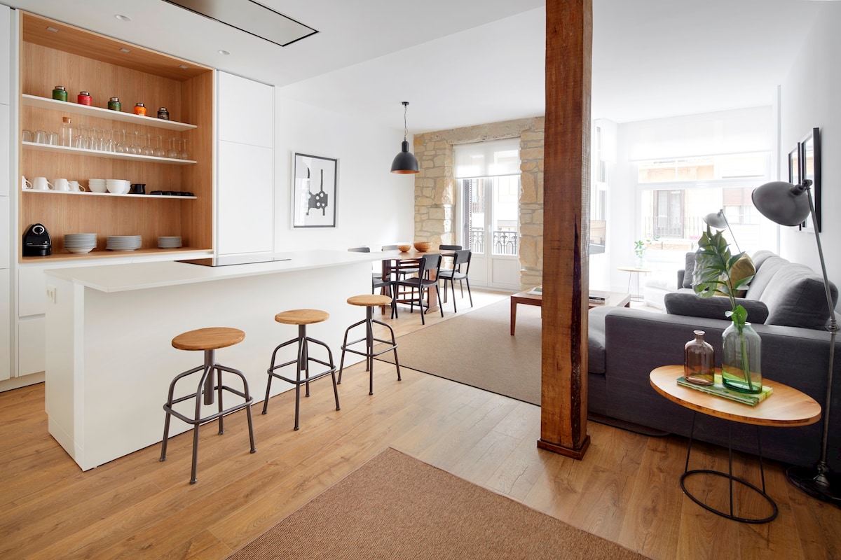 An open-plan living space features a modern kitchen island with three wooden stools. A dining area, with a wooden table and chairs, is visible beside a comfortable gray sofa. Natural light floods in through large windows, enhancing the bright atmosphere of the room.