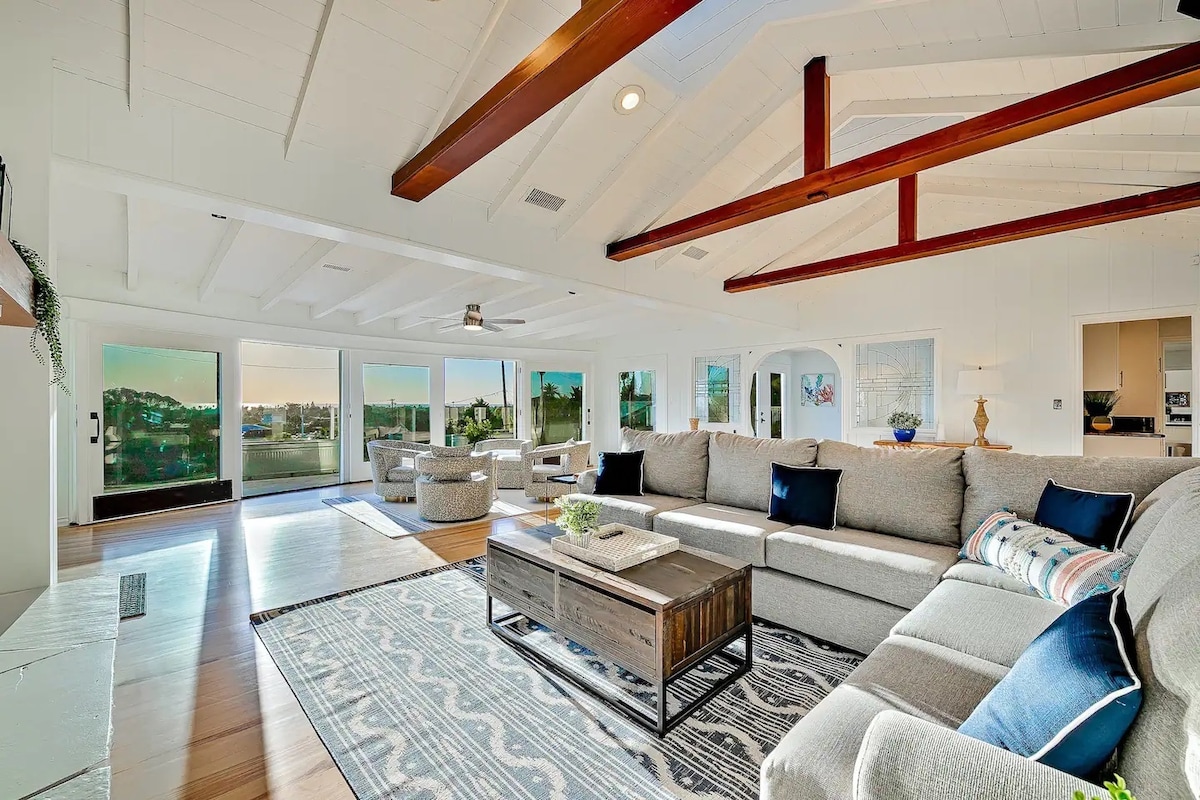 A spacious living room is highlighted by a high, beamed ceiling and large windows that offer expansive views. Comfortable seating arrangements feature neutral upholstery and accent pillows. A wooden coffee table with decorative items anchors the space, complemented by a stylish area rug.