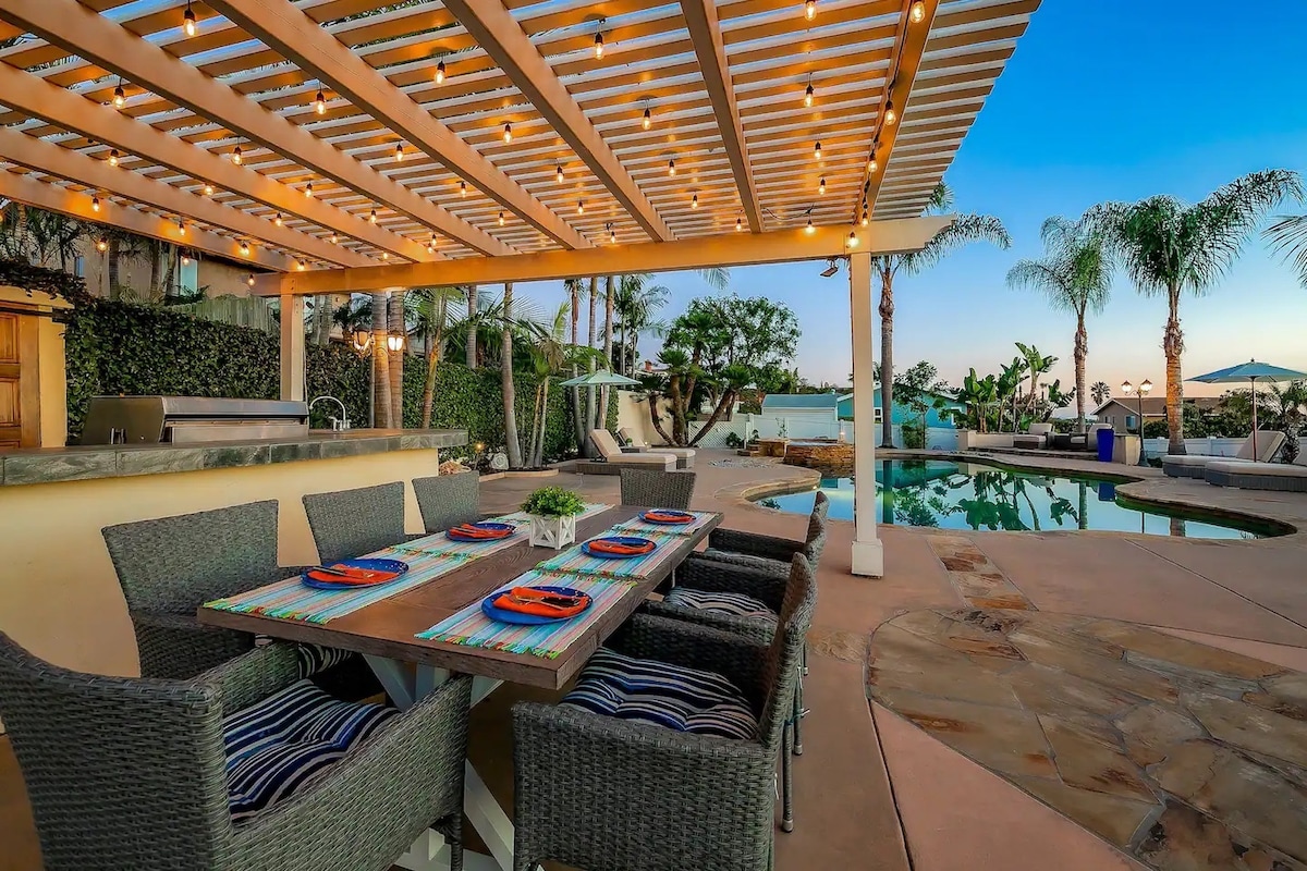 An outdoor dining area is presented under a structured pergola with warm string lights. A large table is surrounded by comfortable, woven chairs. The inviting pool and lounge chairs are visible, complemented by palm trees swaying gently in the breeze.