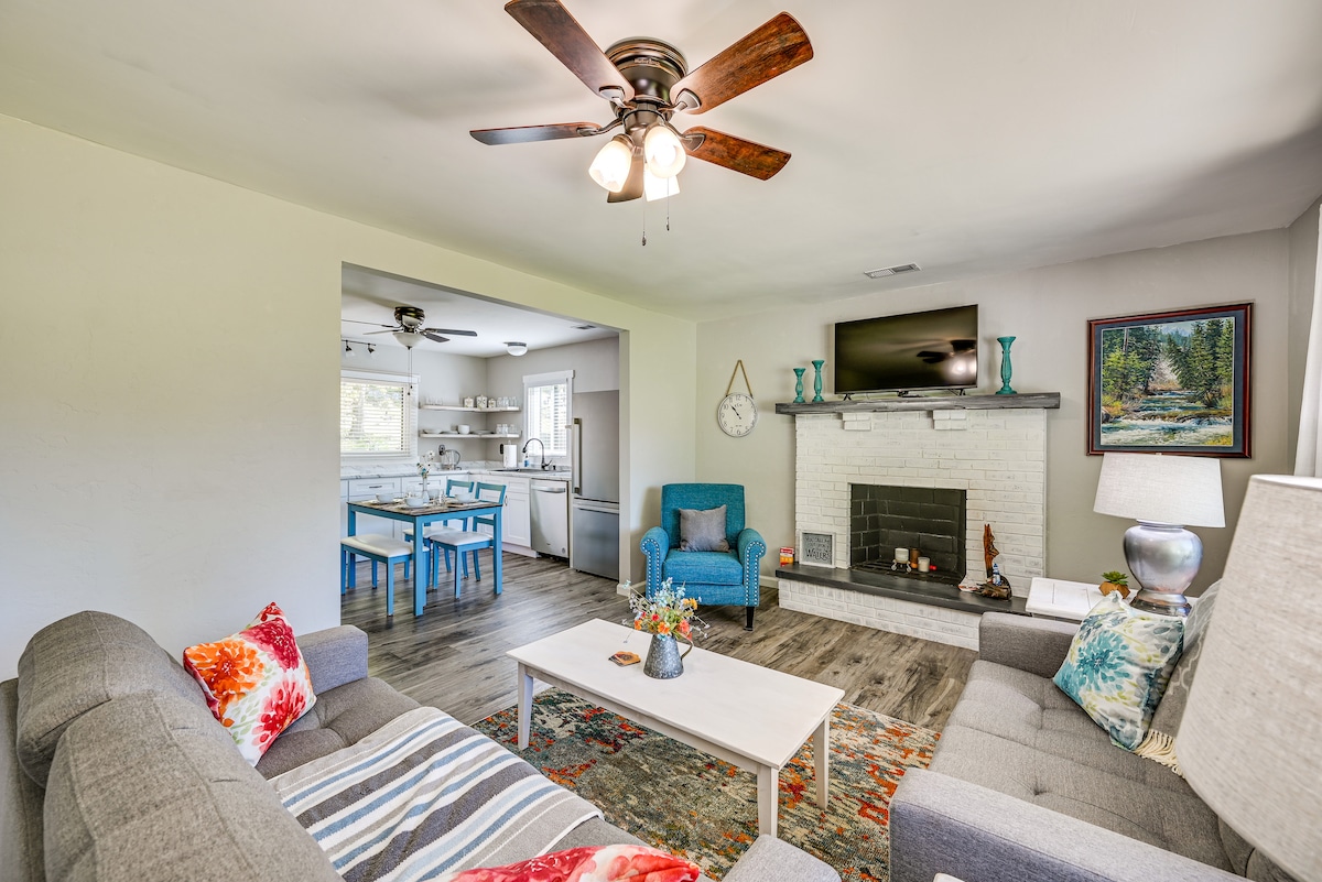 A welcoming living area features a light-colored sofa and matching chairs, with colorful throw pillows adding a touch of vibrancy. A coffee table sits atop an area rug, while a wall-mounted TV and decorative artwork complement the space. The open layout connects to the kitchen area.
