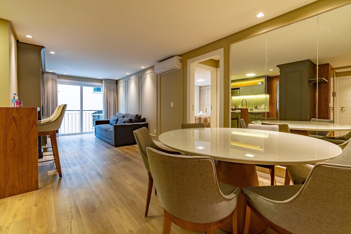An open-plan living area includes a circular dining table surrounded by several chairs. A modern sofa is positioned in the background, with natural light filtering through large windows. Reflective surfaces enhance the sense of space and light in the room.