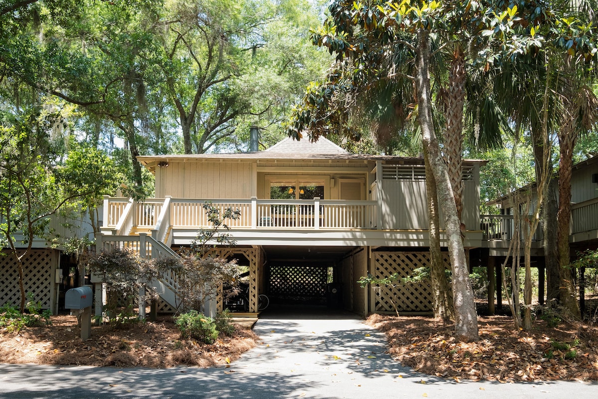 A light-colored cottage is elevated on stilts, surrounded by lush greenery and mature trees. The spacious front porch is accentuated by railing, offering an inviting entry. Underneath, a covered area provides parking space, complemented by simple landscaping and a gentle path leading to the entrance.