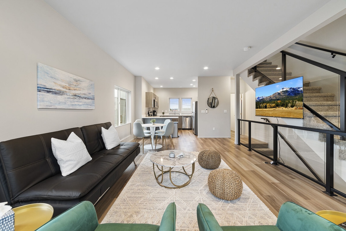 A modern living area showcases a sleek futon sofa with decorative pillows, paired with a round coffee table atop a textured area rug. A dining table for four is positioned nearby, while a large window allows natural light to brighten the space. A smart TV is mounted on the wall.