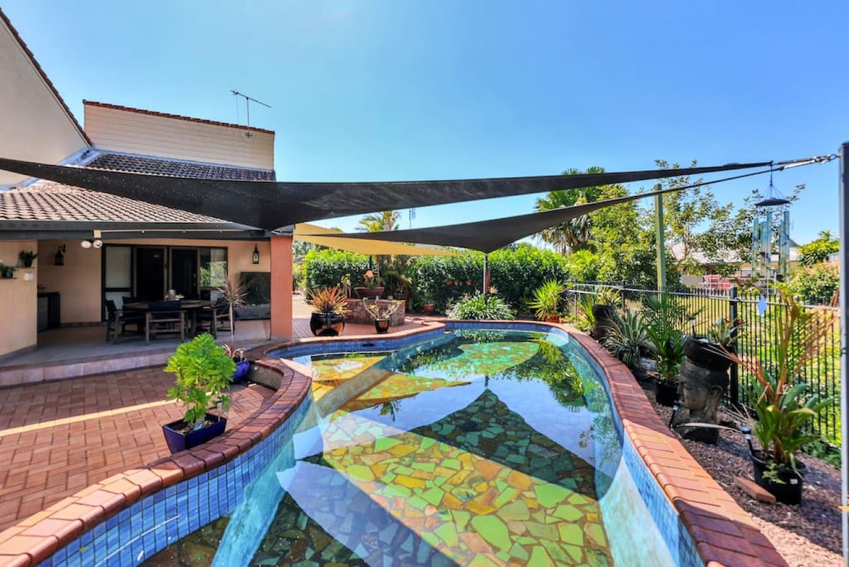 The outdoor area features a curved private pool, adorned with colorful tiles showcasing natural patterns. Shade sails provide protection from direct sunlight. Lush plants border the pool, complemented by a spacious dining area and sliding glass doors that open to the interior.