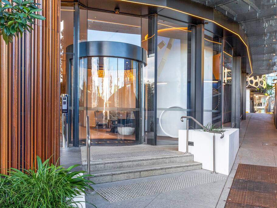 The entrance to the building showcases modern architectural elements, featuring large glass doors framed by warm wooden panels. A gentle slope leads to the entrance, with neatly arranged greenery on either side, creating an inviting and accessible access point.