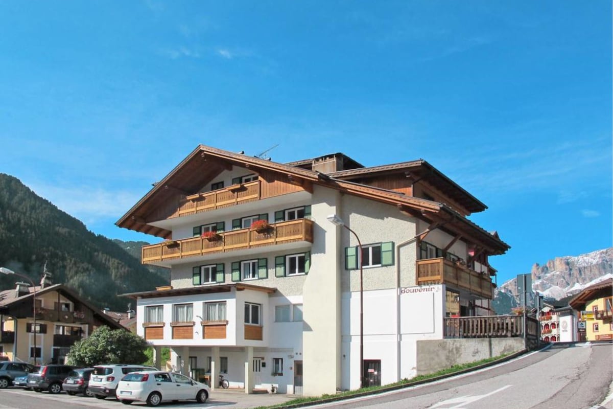 The three-story building is nestled against a mountainous backdrop, featuring a wooden facade with multiple balconies. Car parking spaces are visible at the front, and the surrounding area showcases a blend of greenery and distant peaks under a clear blue sky.