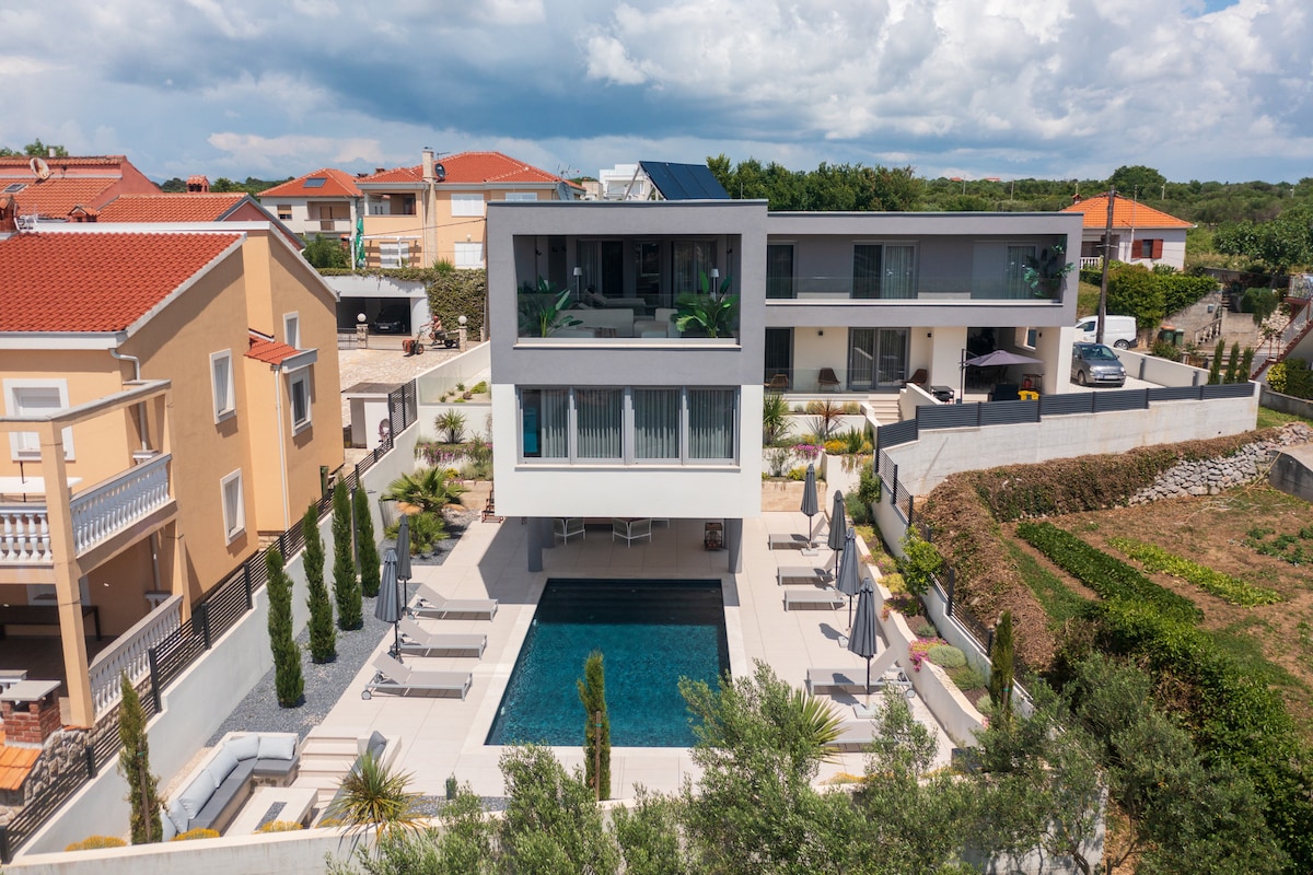 The villa is set on a spacious property, featuring a sleek, modern design with multiple levels. A private pool is surrounded by loungers, while landscaped greenery and tall cypress trees provide a serene environment. Nearby residential buildings are visible, framed by a clear sky.