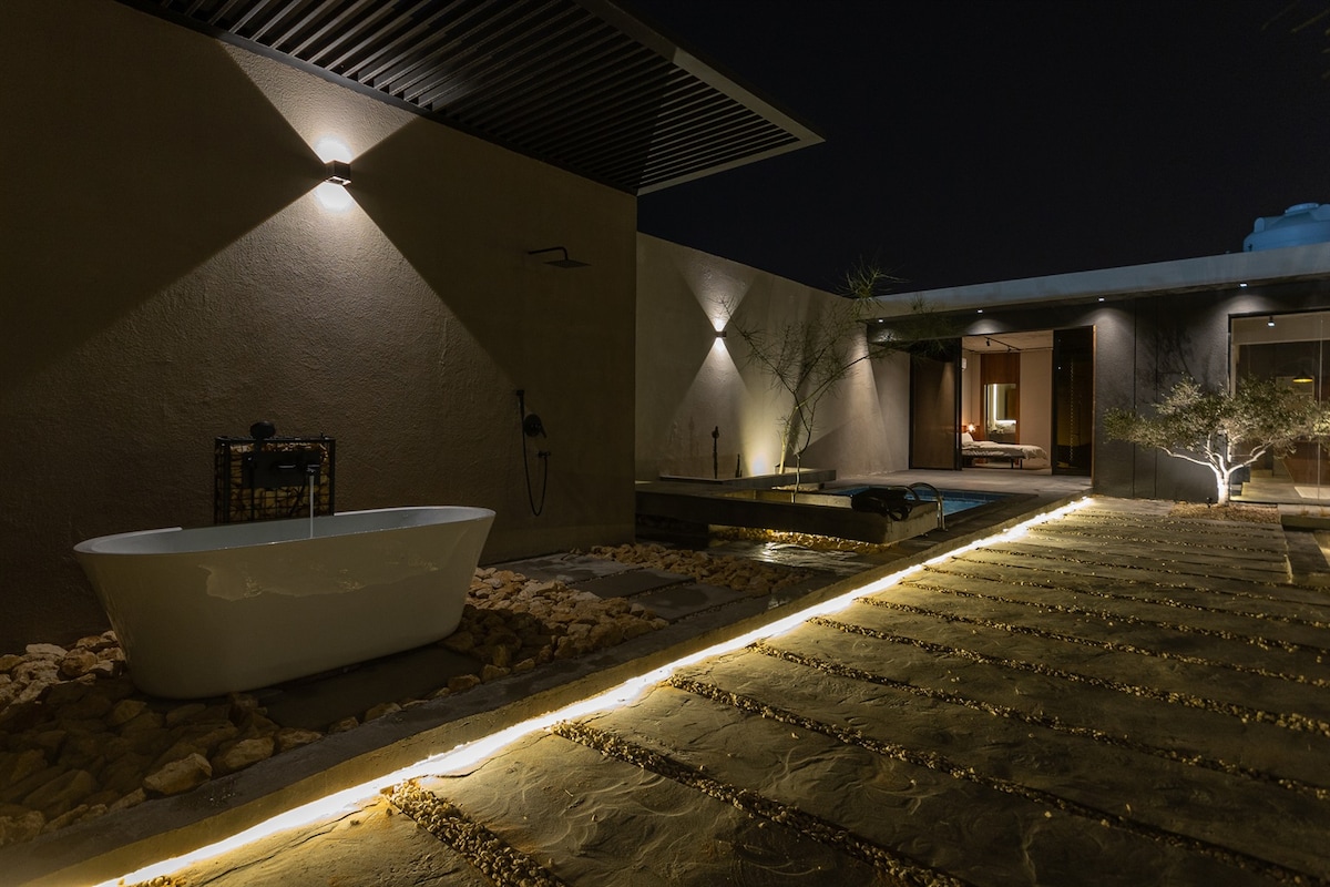 An outdoor space illuminated at night features a freestanding bathtub alongside a tranquil pathway. Subtle lighting enhances the natural stone tiles and desert landscaping, leading to an elegant entrance where indoor and outdoor areas seamlessly connect.