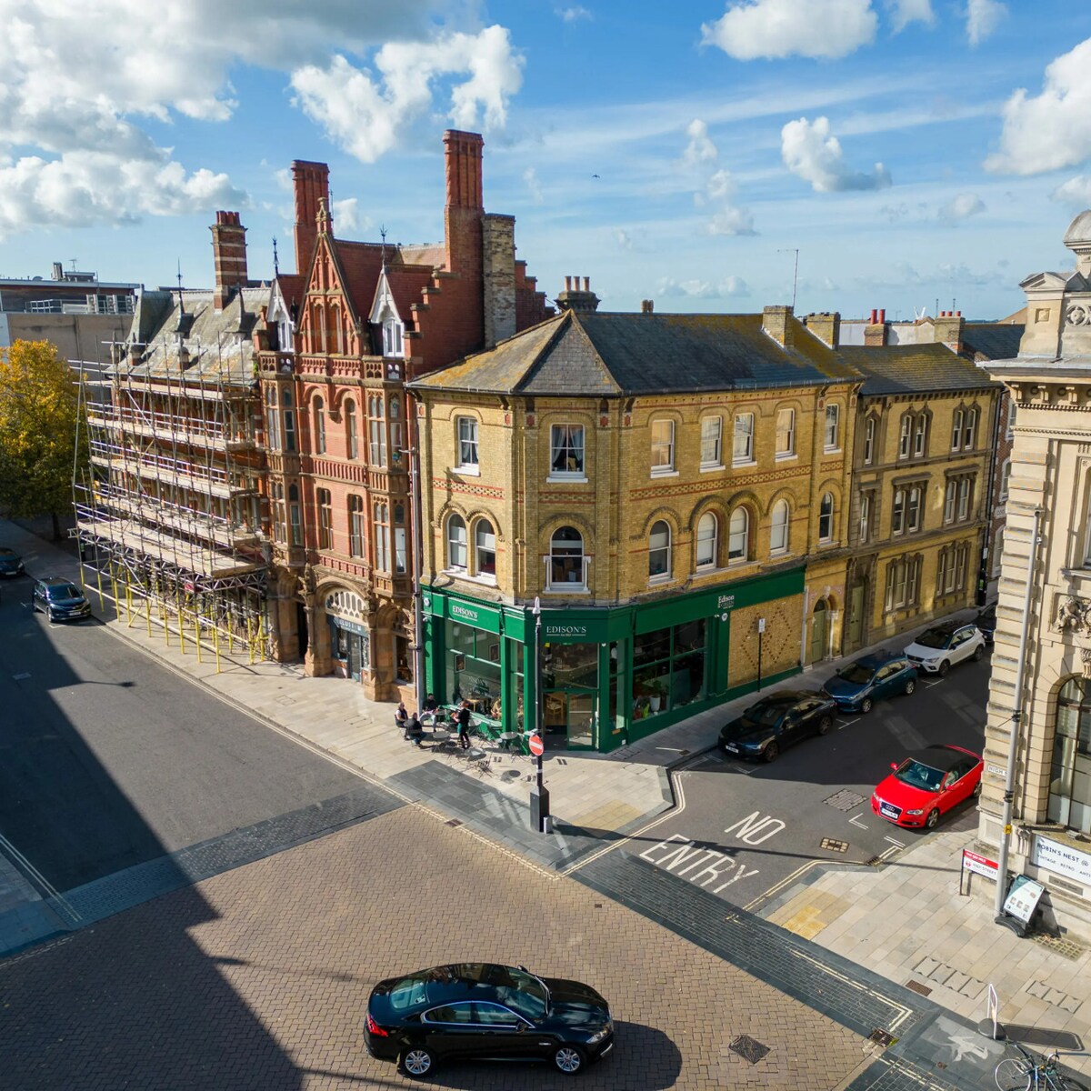 The image captures a view of a bustling street corner, showcasing a mix of historic and modern architecture. The scene includes buildings with intricate facades and large windows, alongside parked cars and street signage. The sky is partly cloudy, providing a dynamic backdrop.
