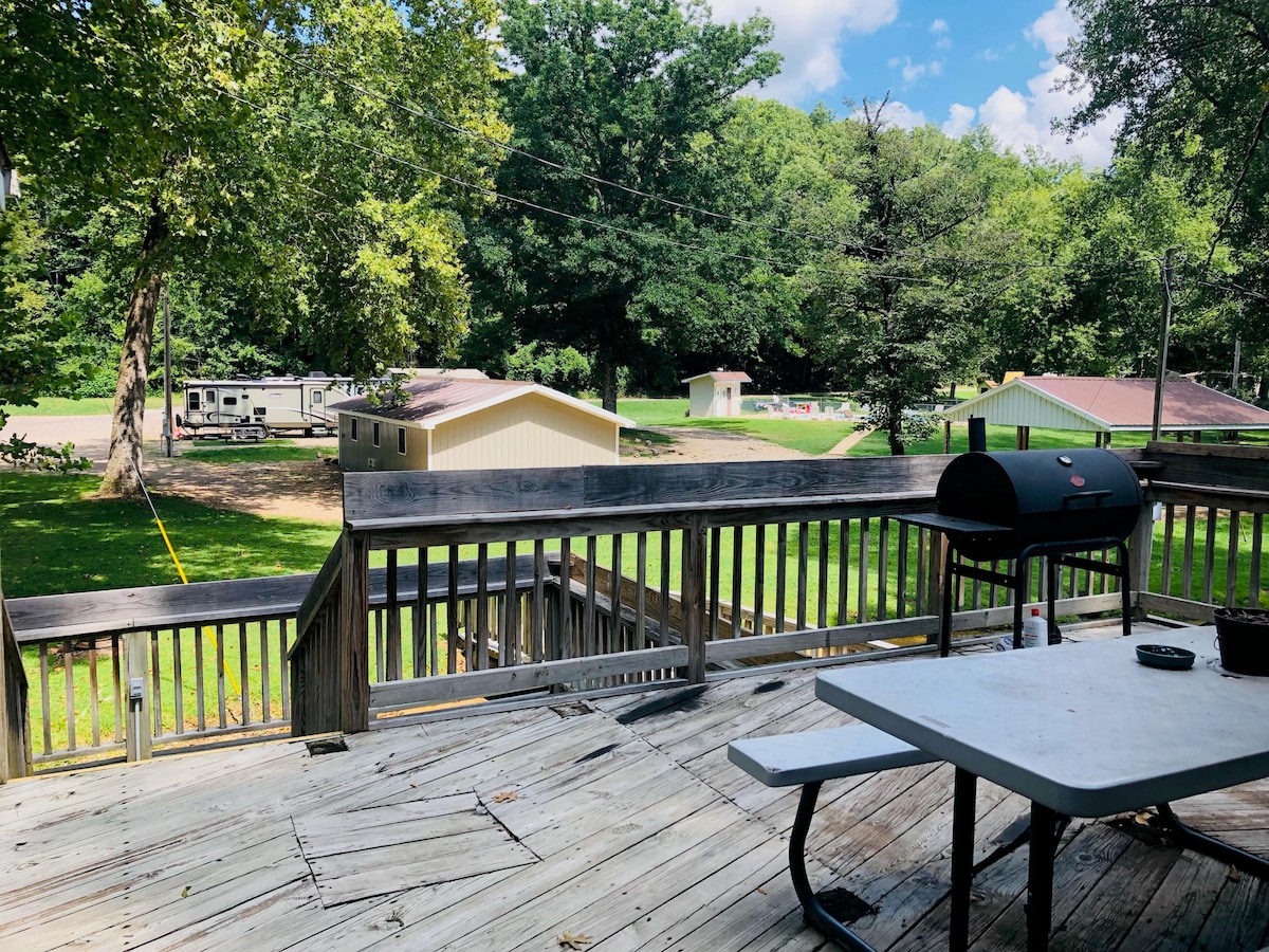 A wooden deck provides an outdoor area for relaxation, featuring a charcoal grill and a table. Green trees create a natural backdrop, while a grassy area extends beyond the deck, which includes structures in the distance.