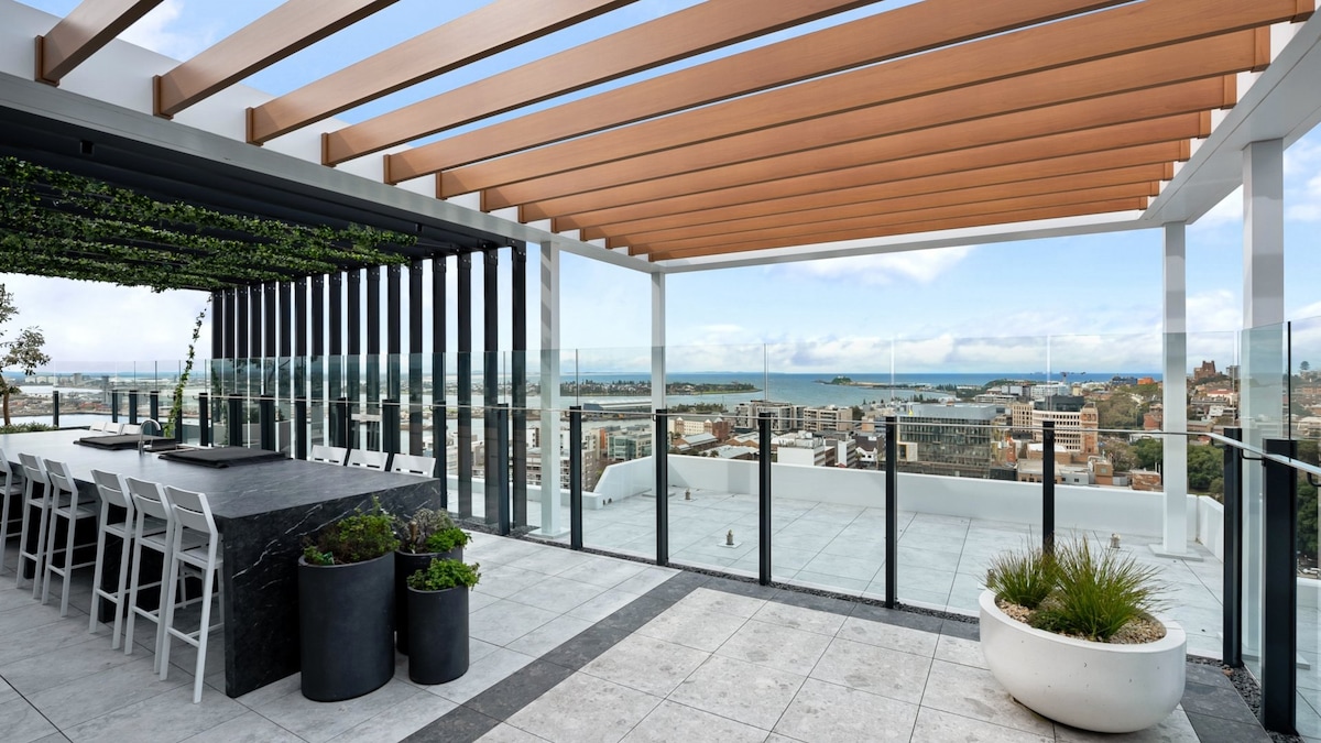 A rooftop terrace features outdoor dining with a large stone table surrounded by white chairs. Lush greenery is integrated into the design, and a glass railing provides an unobstructed view of the harbor and skyline. The space is illuminated by natural light.