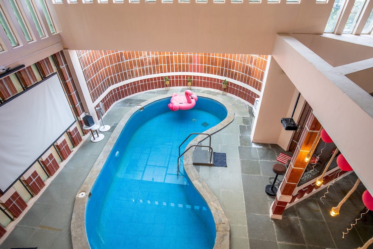 An indoor private pool is showcased, framed by elegant walls complemented by large windows allowing ample natural light. A pink inflatable float adds a playful touch to the serene blue water. A staircase leads to additional amenities, enhancing the inviting atmosphere of the space.