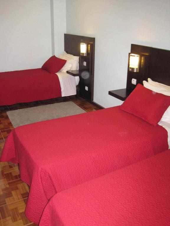 A bright sleeping area features two beds with red covers and matching decorative pillows. Each bed is positioned against a dark headboard with wall-mounted lamps for reading. A neutral carpet is placed centrally, enhancing the space's warmth.