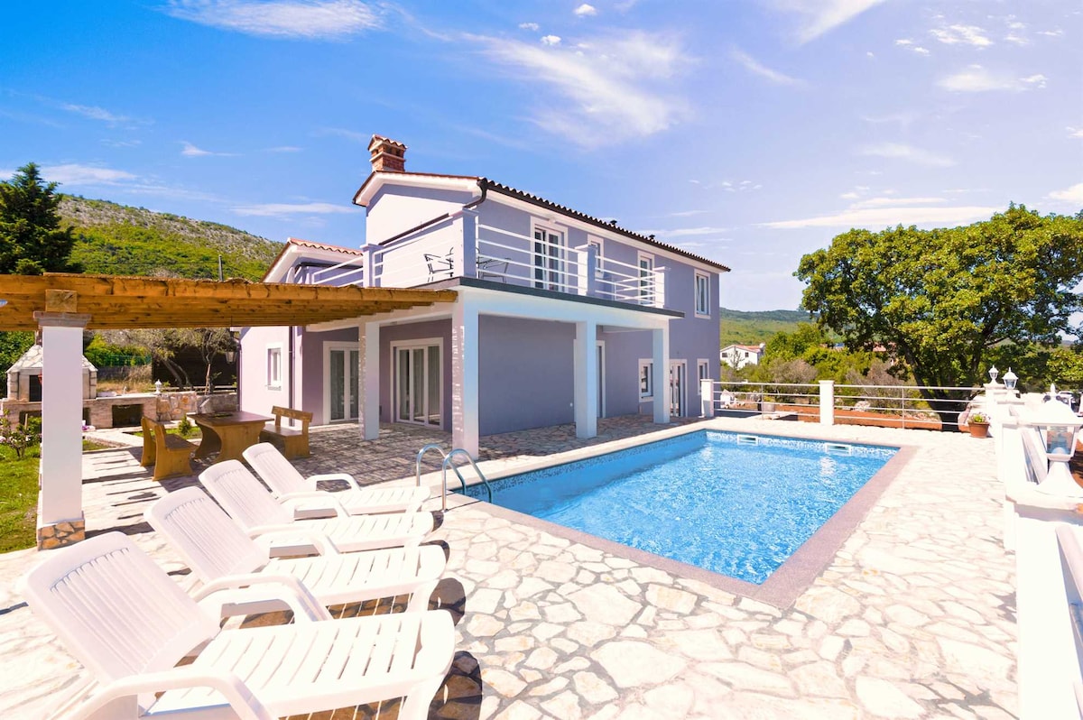 A spacious villa exterior is shown, featuring a private pool surrounded by stone paving. Sun loungers are positioned by the pool. The two-story structure presents a modern facade with a balcony, framed by green trees and distant hills under a clear blue sky.