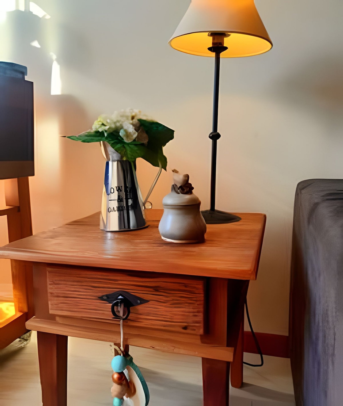 A wooden bedside table is depicted, featuring a drawer and adorned with a small decorative charm. A metallic vase with flowers adds a touch of nature, while a lamp stands beside it, casting a warm glow in the corner of the room.