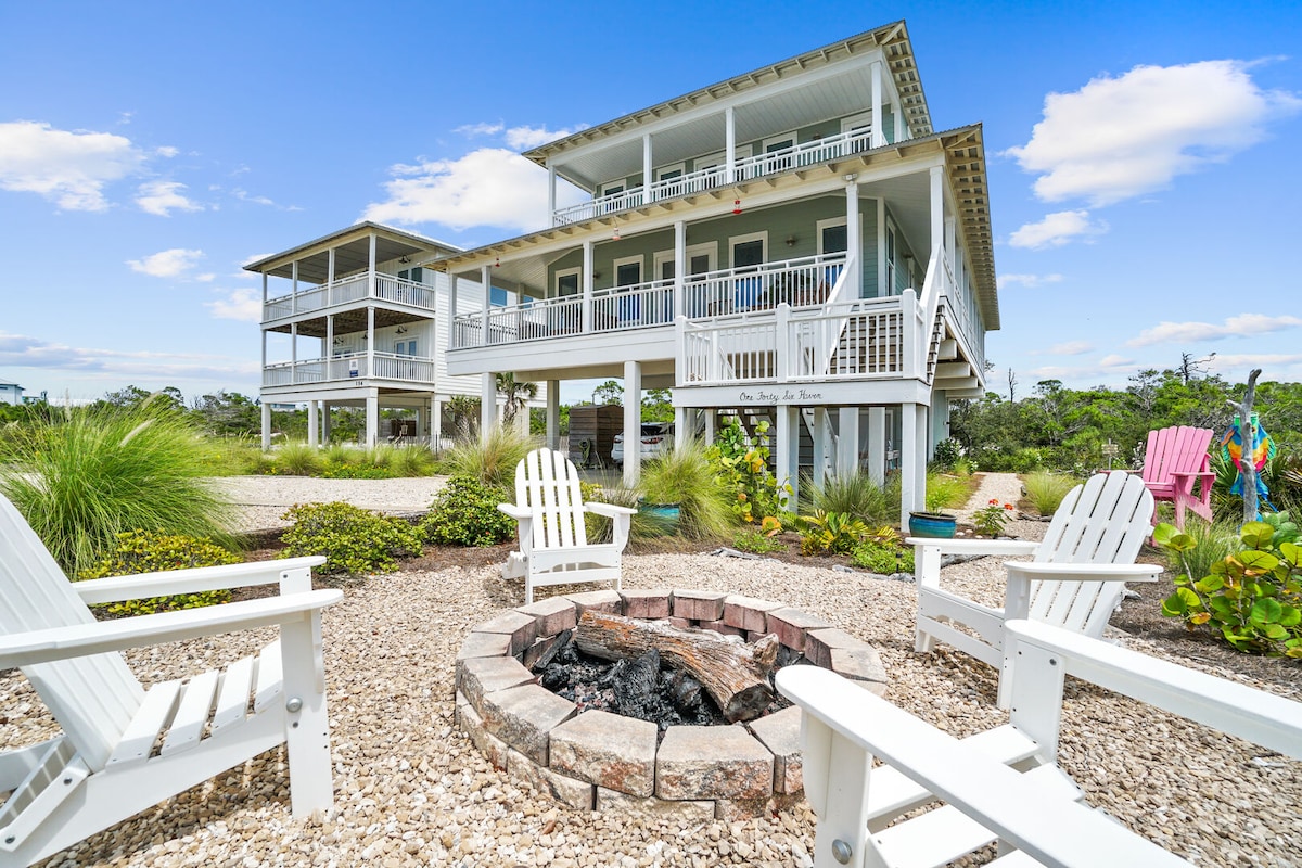 The exterior of the multi-level home is shown, emphasizing its spacious balconies and inviting design. Surrounding the fire pit are several white Adirondack chairs. Lush greenery and landscaping enhance the outdoor atmosphere, creating a welcoming environment for relaxation.