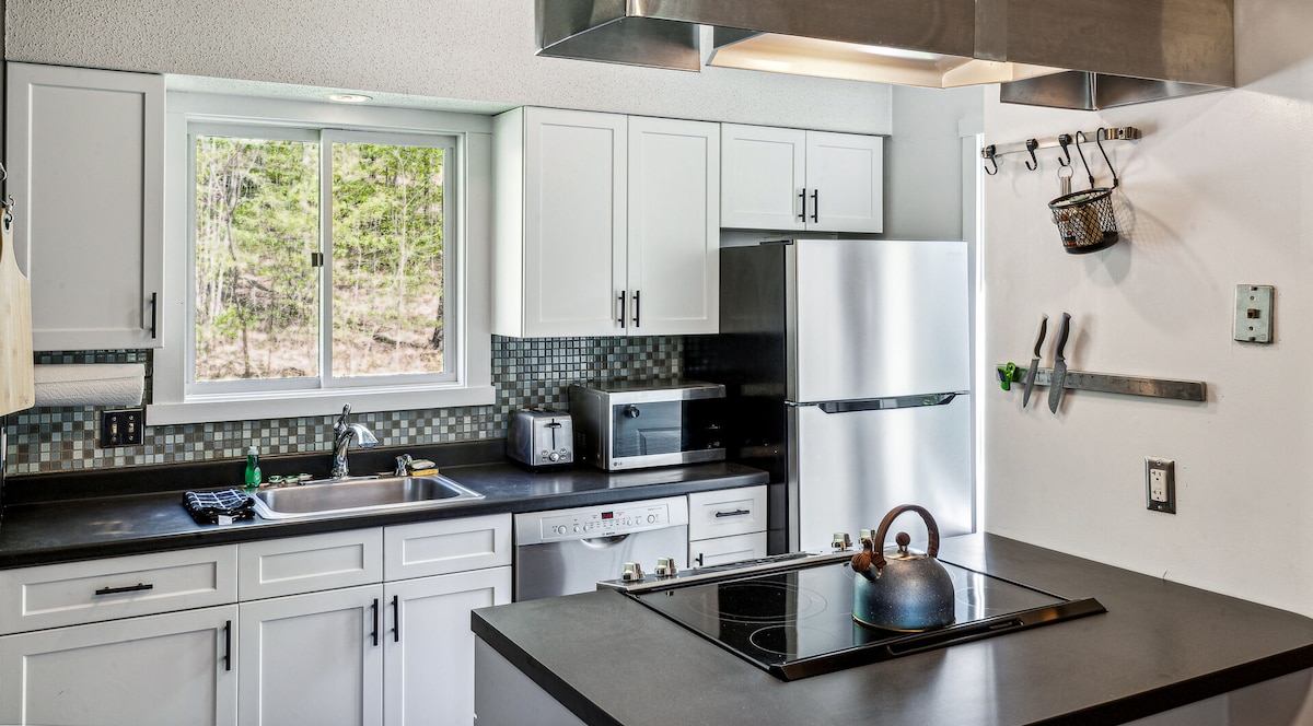 The updated kitchen features white cabinetry and a black countertop. Stainless steel appliances, including a refrigerator and dishwasher, are prominently displayed. A window offers natural light, overlooking a green landscape. Cooking equipment is neatly arranged, enhancing functionality in this modern space.