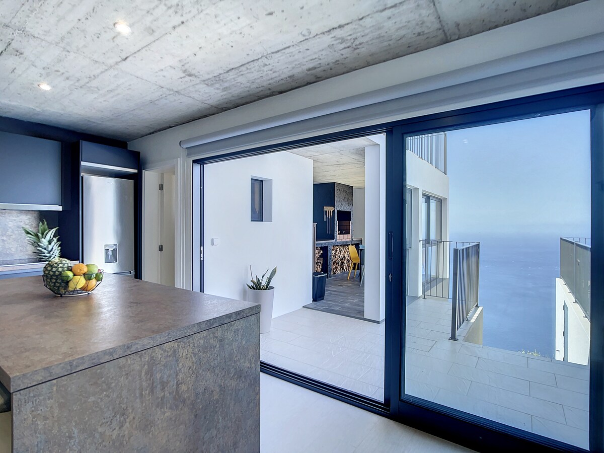 A modern kitchen is seen, featuring a large island with a fruit bowl at its center. Expansive glass doors lead to an outdoor area, showcasing stunning ocean views. The minimalist design is complemented by sleek cabinetry and a fresh ambiance.