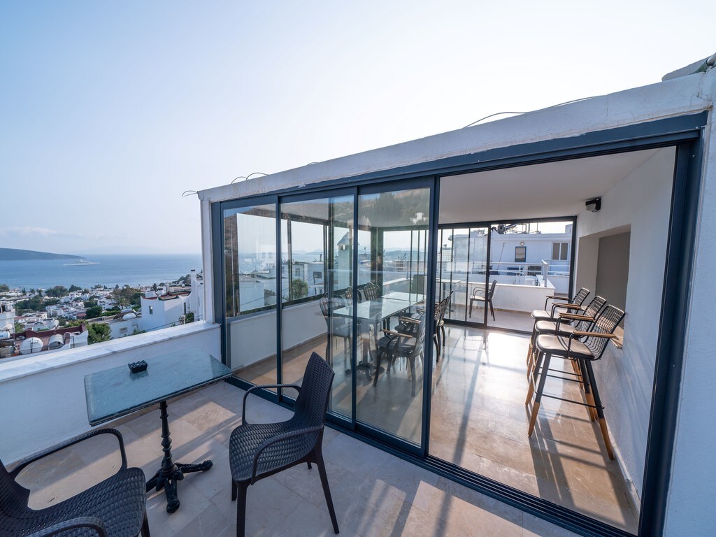 A spacious communal terrace features large glass doors opening to a panoramic view of the surrounding area. Outdoor seating includes several high-backed chairs and a table, creating a welcoming space for relaxation or work while enjoying the sea and town views.