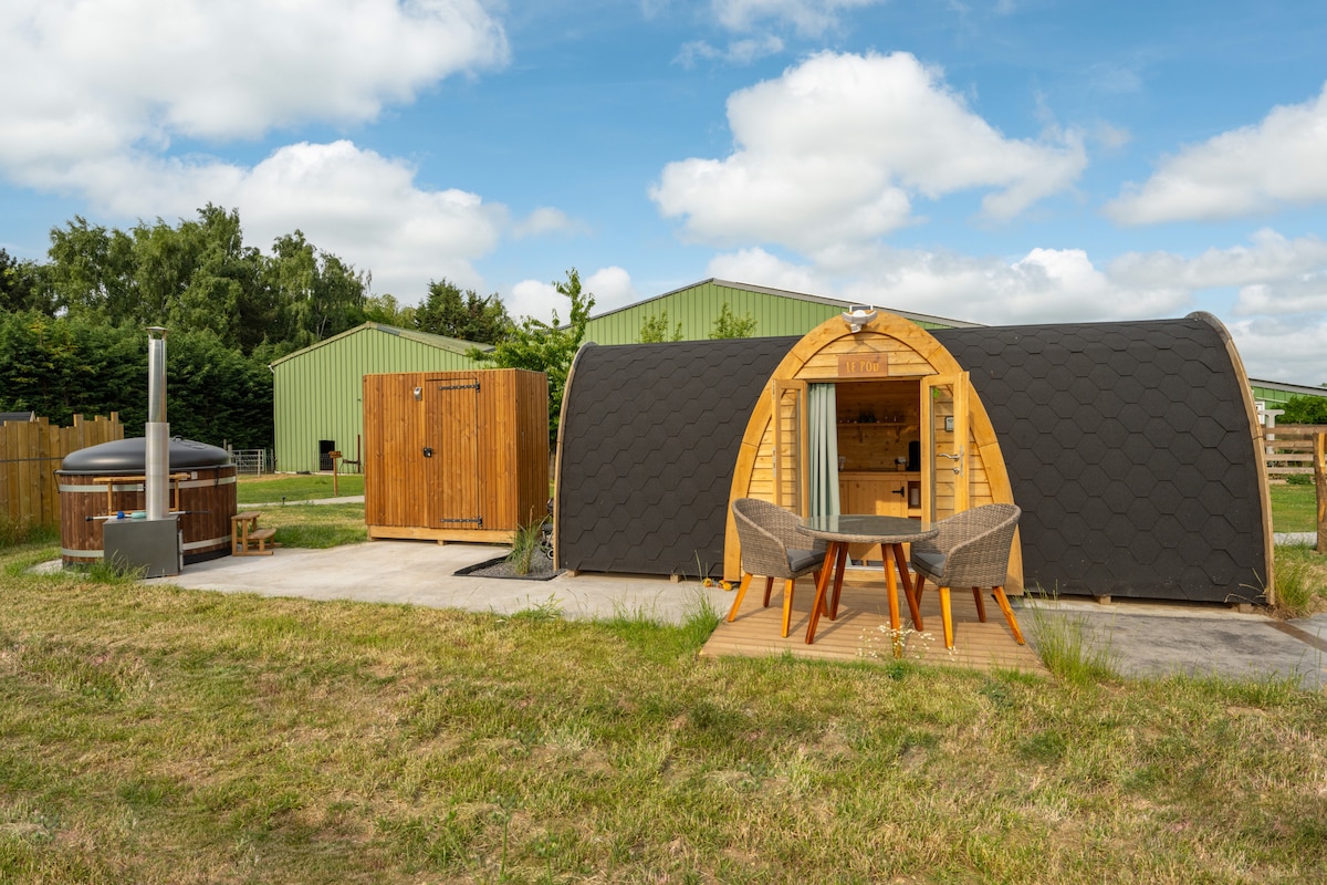 A cozy outdoor pod is shown with a round wooden hot tub nearby. A wooden deck features a small table and two chairs, placed on a grassy area. The background includes structures with green roofs, surrounded by trees under a partly cloudy sky.