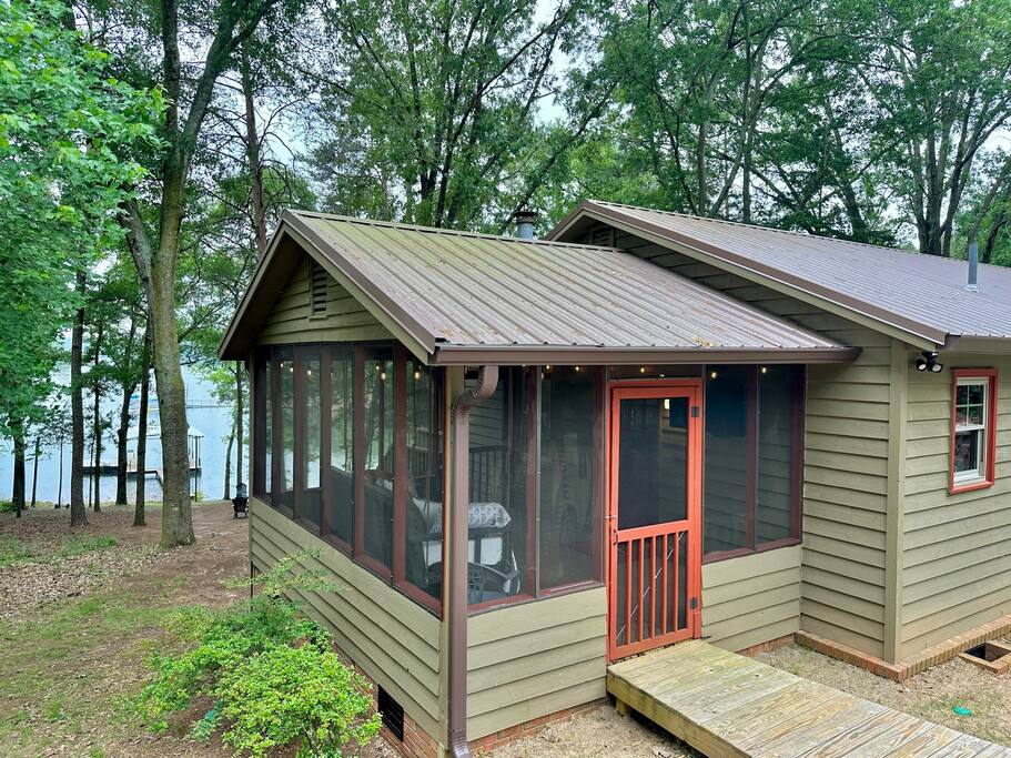 The exterior of a cozy house is framed by green trees, providing a calming natural backdrop. A screened-in porch with wooden steps leads to a pathway. The structure features a peaked roof and blends seamlessly with its surroundings, showcasing a quiet lakeside atmosphere.