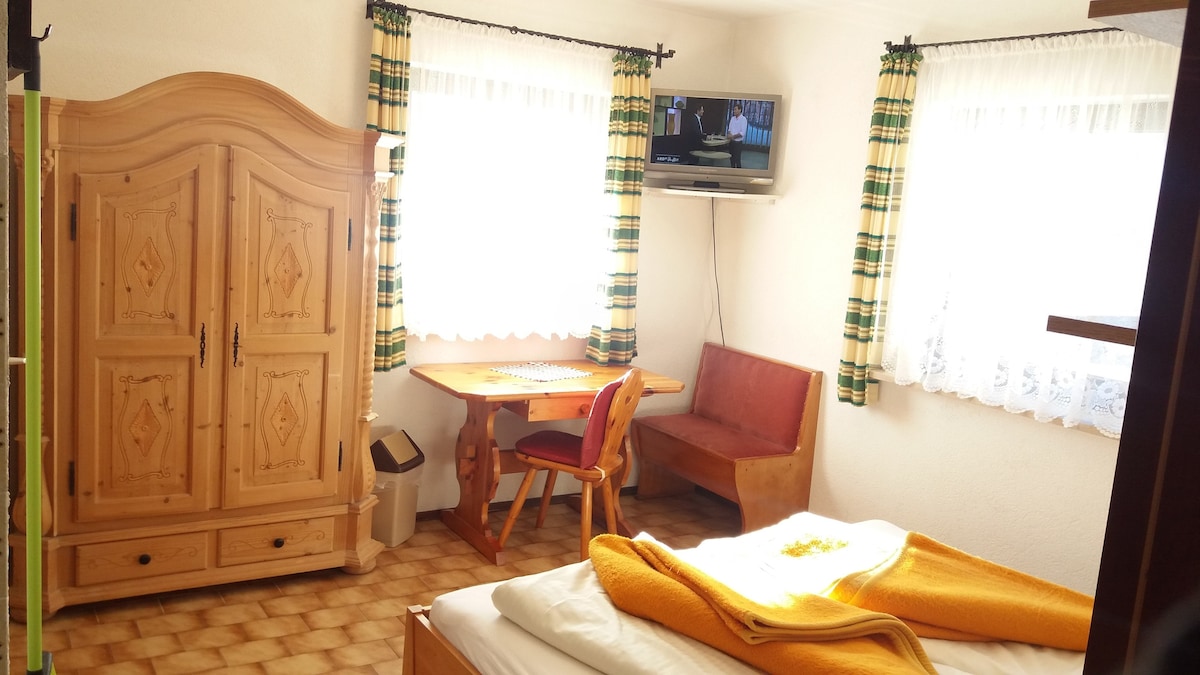 A bright room is presented with natural light filtering through two large windows. A cozy sleeping area features an unmade bed with a vibrant orange blanket. A wooden table with two chairs and a small seating area are included. A TV is mounted on the wall.