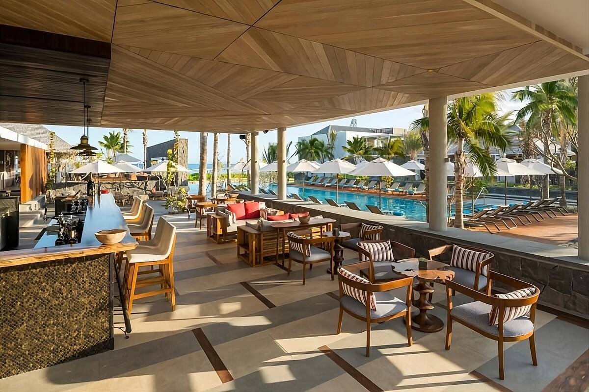 An outdoor dining area features a stylish bar with high-backed chairs, and multiple tables arranged for seating. Umbrellas provide shade over the loungers by the pool, creating a relaxed atmosphere amidst palm trees and tranquil waters.
