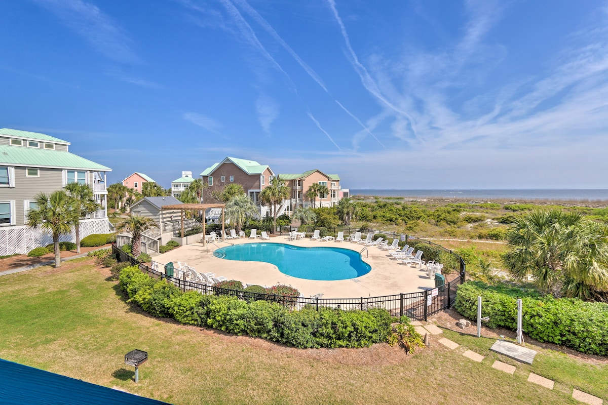 An outdoor pool is framed by lush greenery and palm trees, with a spacious deck and lounge chairs positioned around it. The surrounding buildings feature colorful exteriors, with blue skies and wispy clouds overhead, providing a bright and inviting atmosphere.