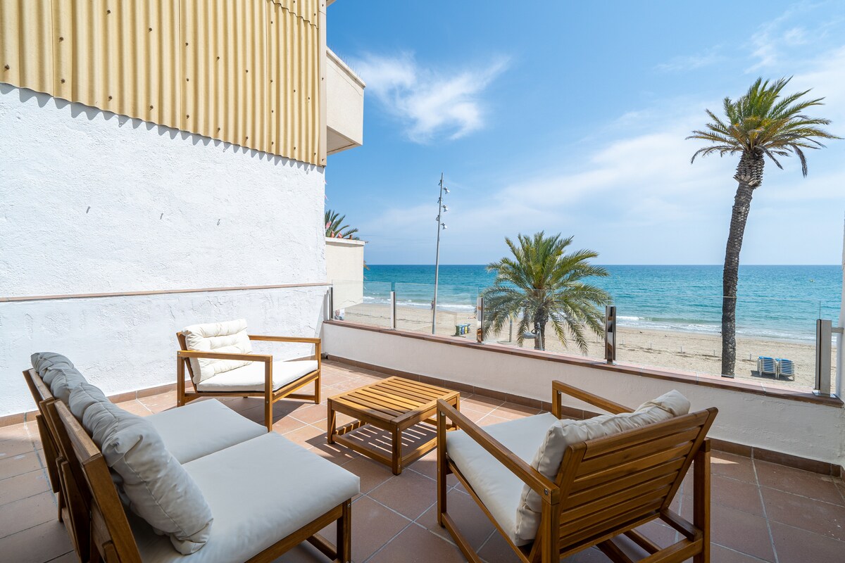 A spacious terrace features several wooden seating options, including a coffee table and cushioned chairs. Framed by palm trees, the area offers unobstructed views of the sandy beach and the sea, with a clear blue sky visible above.