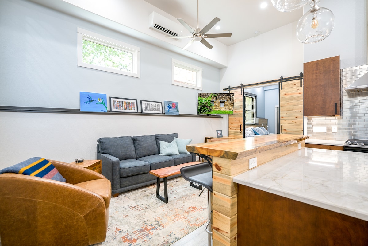 A cozy living area is highlighted by a modern couch and a wooden coffee table. A mounted television on a wooden accent wall complements the space, while a rustic dining counter with stylish bar stools offers additional seating. Natural light filters through large windows.