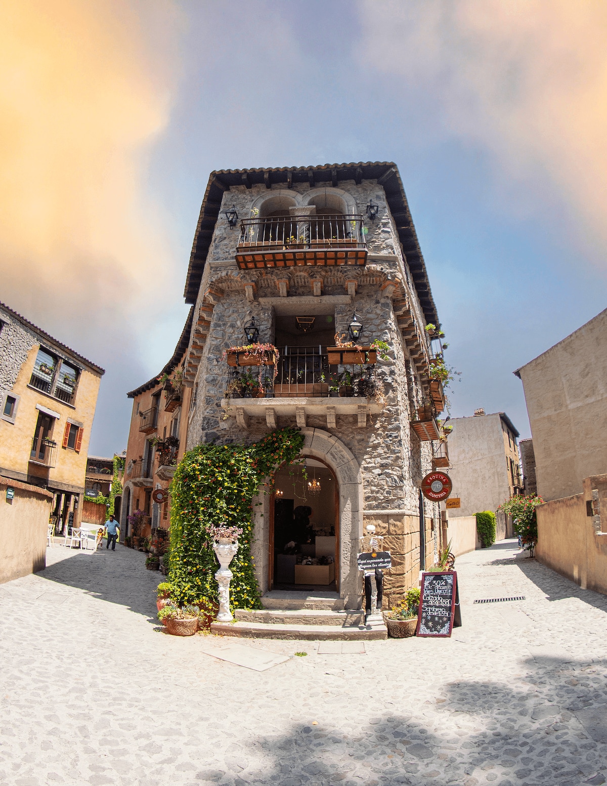 A charming stone building stands along a cobblestone path, featuring intricate architectural details and balconies adorned with greenery. Baskets of flowers hang from the walls, enhancing the picturesque setting. The scene evokes a quaint village atmosphere with nearby structures visible in the background.
