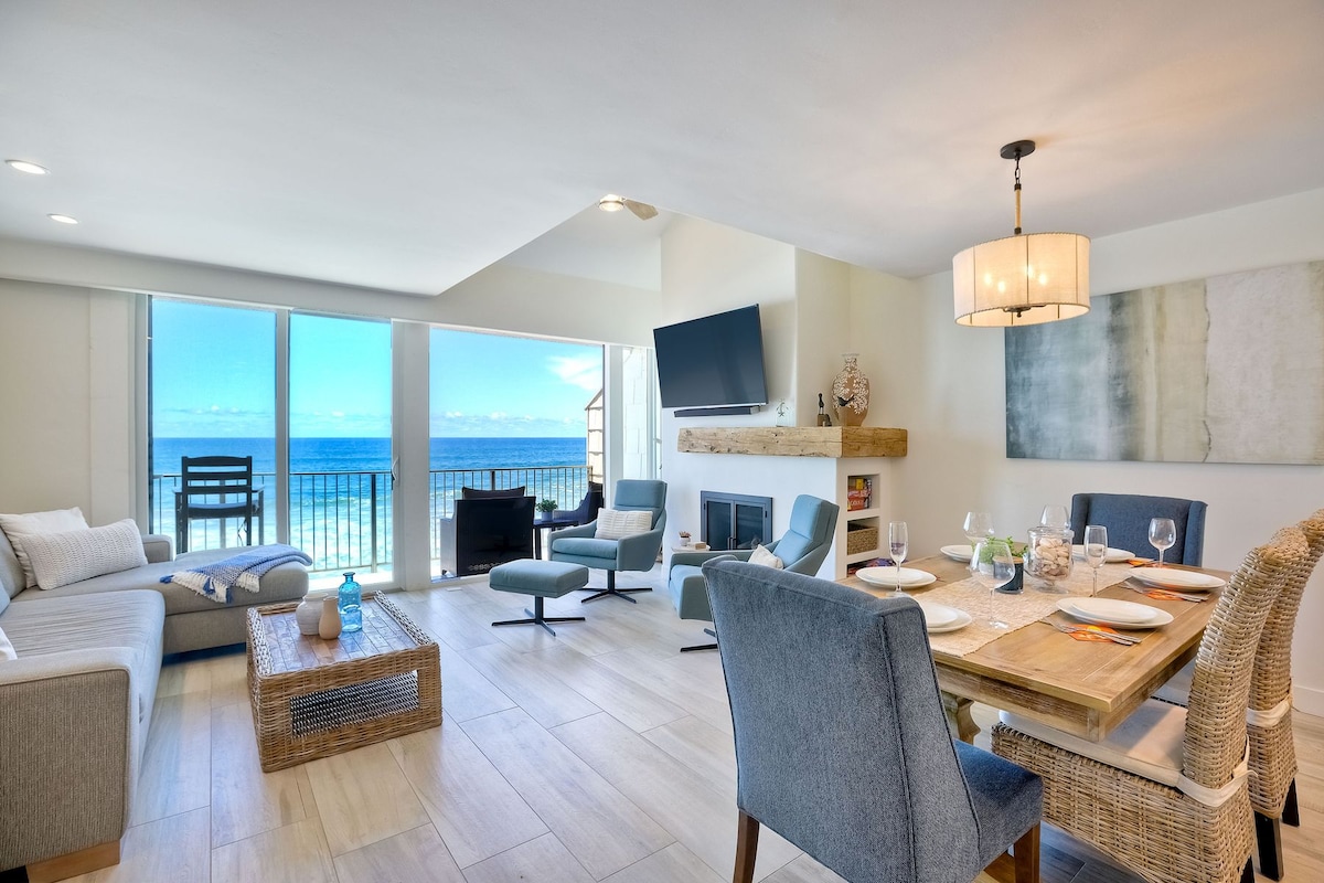 An open-concept living area features modern furnishings and a dining table set for six. Large glass doors provide unobstructed views of the ocean. A comfortable seating arrangement includes a sectional couch and stylish chairs, creating a welcoming atmosphere for gatherings.