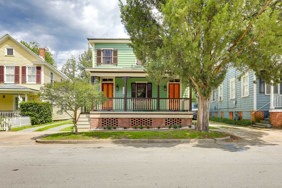 A two-story Victorian-era home is surrounded by greenery, featuring a welcoming covered porch with wooden railings and two front doors. The exterior showcases a light green color with brick detailing, while nearby trees provide shade. Sidewalks lead to the property, enhancing accessibility.
