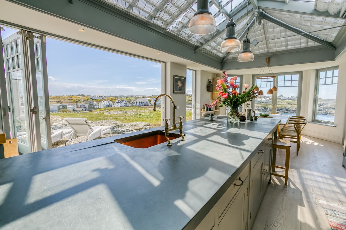 A bright kitchen features a spacious island with a concrete countertop and bar stools. Large bi-fold doors open to reveal views of the landscape beyond, with sunlight illuminating vibrant flowers on the countertop. Distinctive pendant lights hang from the ceiling, enhancing the inviting atmosphere.