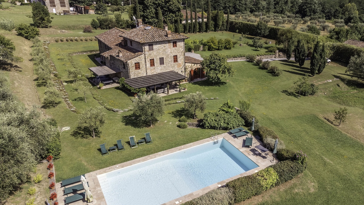 Aerial view of Villa Venere 12 showcases the property surrounded by lush greenery, olive groves, and manicured gardens. The 7x14 metre swimming pool is visible, framed by sun loungers. The villa's rustic stone exterior adds character to the serene landscape.