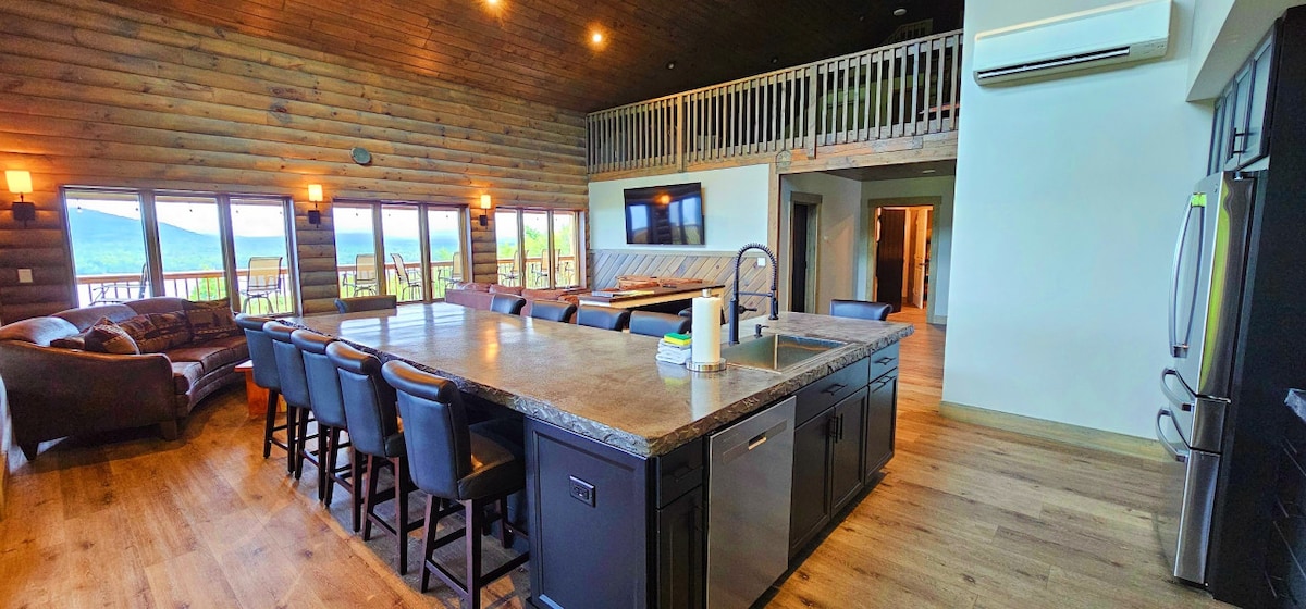 A spacious kitchen with an island featuring seating for eight is highlighted. The room showcases a warm wooden ceiling and large windows that frame views of the surrounding landscape. Comfortable seating is arranged nearby, creating an inviting communal space.