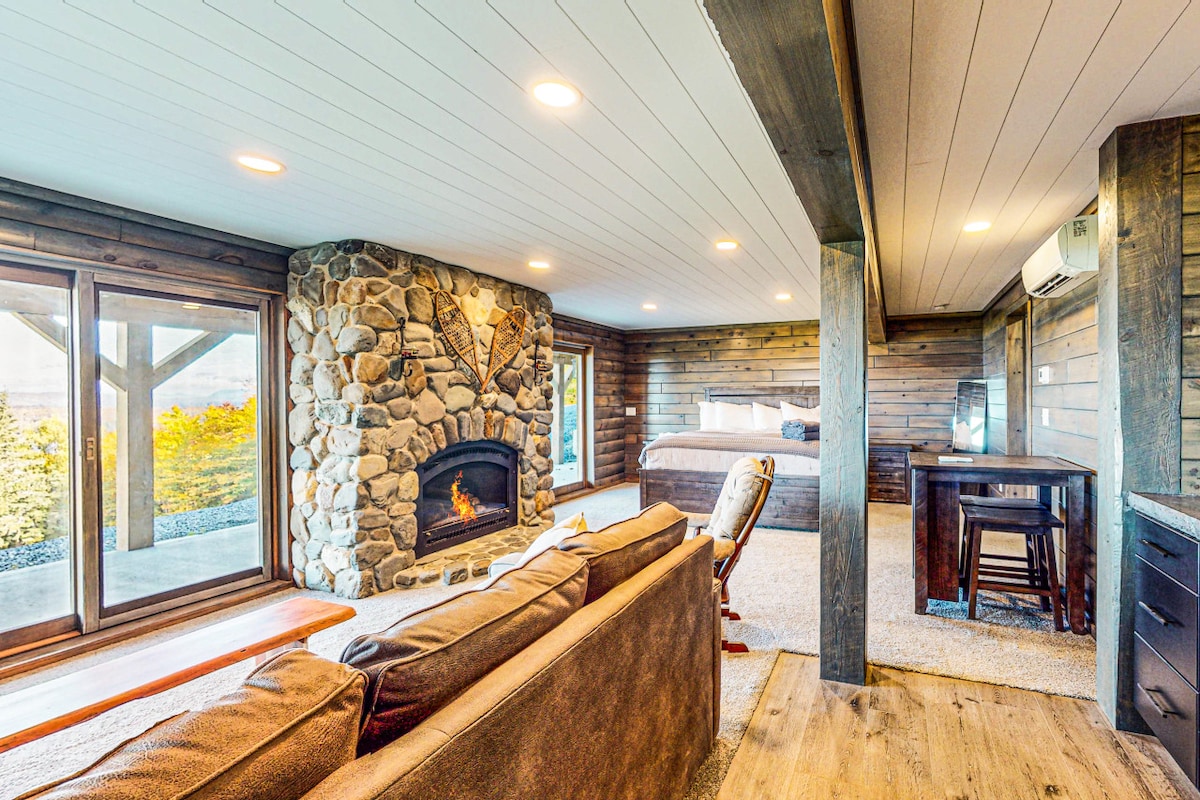 A spacious living area features a stone fireplace as the focal point. Natural light floods in through large windows, framing scenic views. A cozy sofa and a small dining table create a welcoming space, while a plush bed is visible in the background, enhancing the room's comfort.