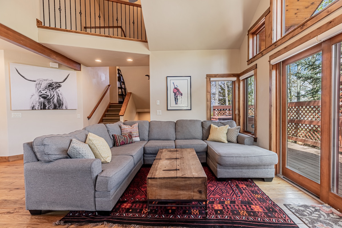 A spacious living area features a large gray sectional sofa adorned with mixed-patterned cushions. A wooden coffee table sits centrally, complemented by a decorative rug. Bright natural light fills the room through expansive windows, highlighting the rustic charm of the wooden accents and artwork on the walls.