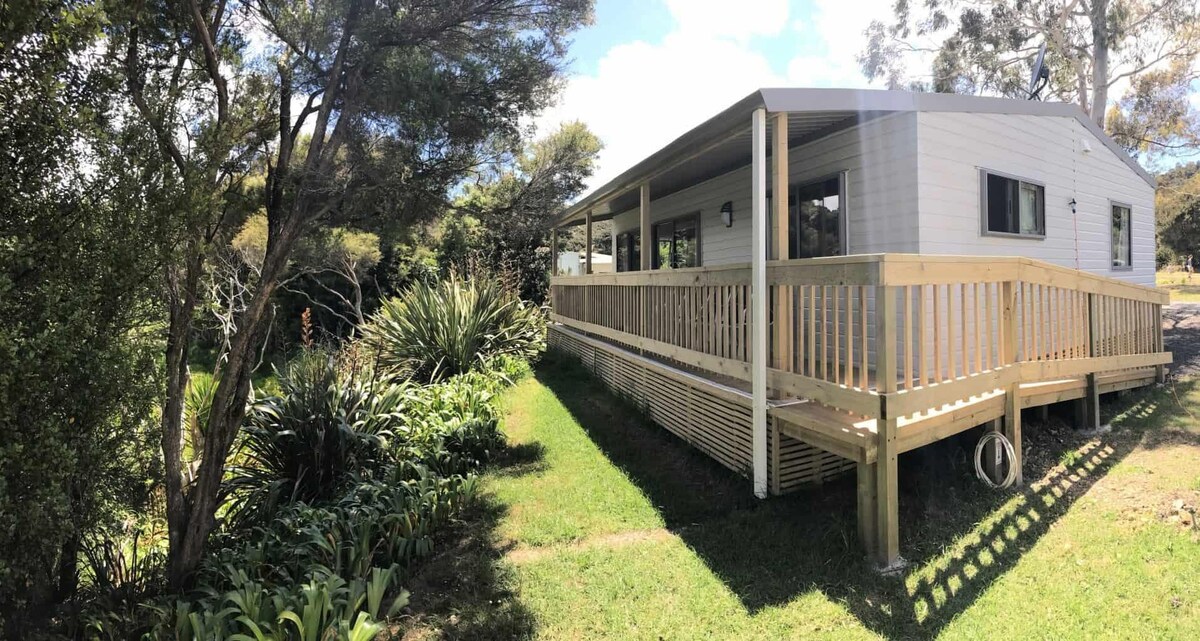 The exterior of the bungalow is shown, featuring a wraparound deck with wooden railings. Lush greenery and plants line the ground, complementing the natural surroundings. The structure has large windows, allowing for ample natural light while blending with the landscape.