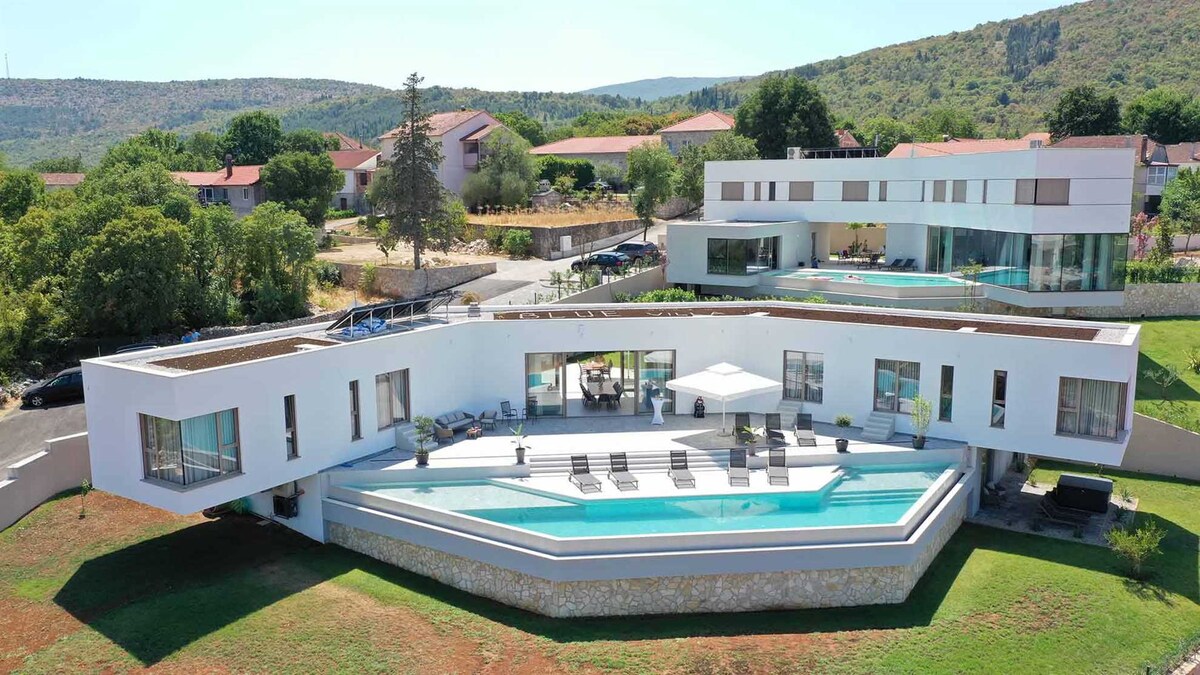 A modern villa is prominently displayed, featuring geometric architecture and expansive glass windows. An outdoor pool surrounded by deck chairs complements a large terrace that offers scenic views of the surrounding green landscape and neighboring buildings.