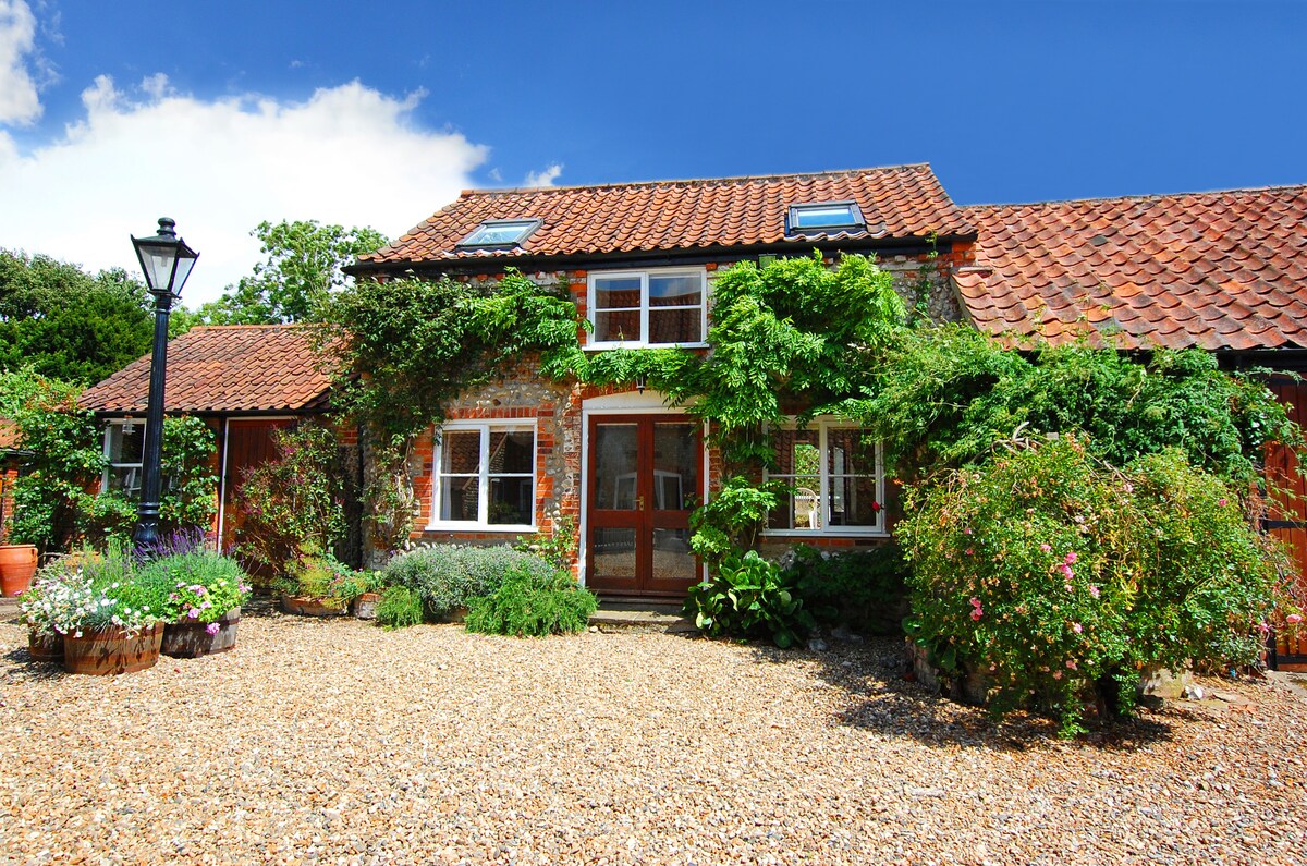 A charming cottage exterior is framed by colorful flower beds and lush greenery. The structure features a tiled roof and multiple windows, inviting natural light. A gravel pathway leads to the entrance, enhancing the cottage's quaint appeal.