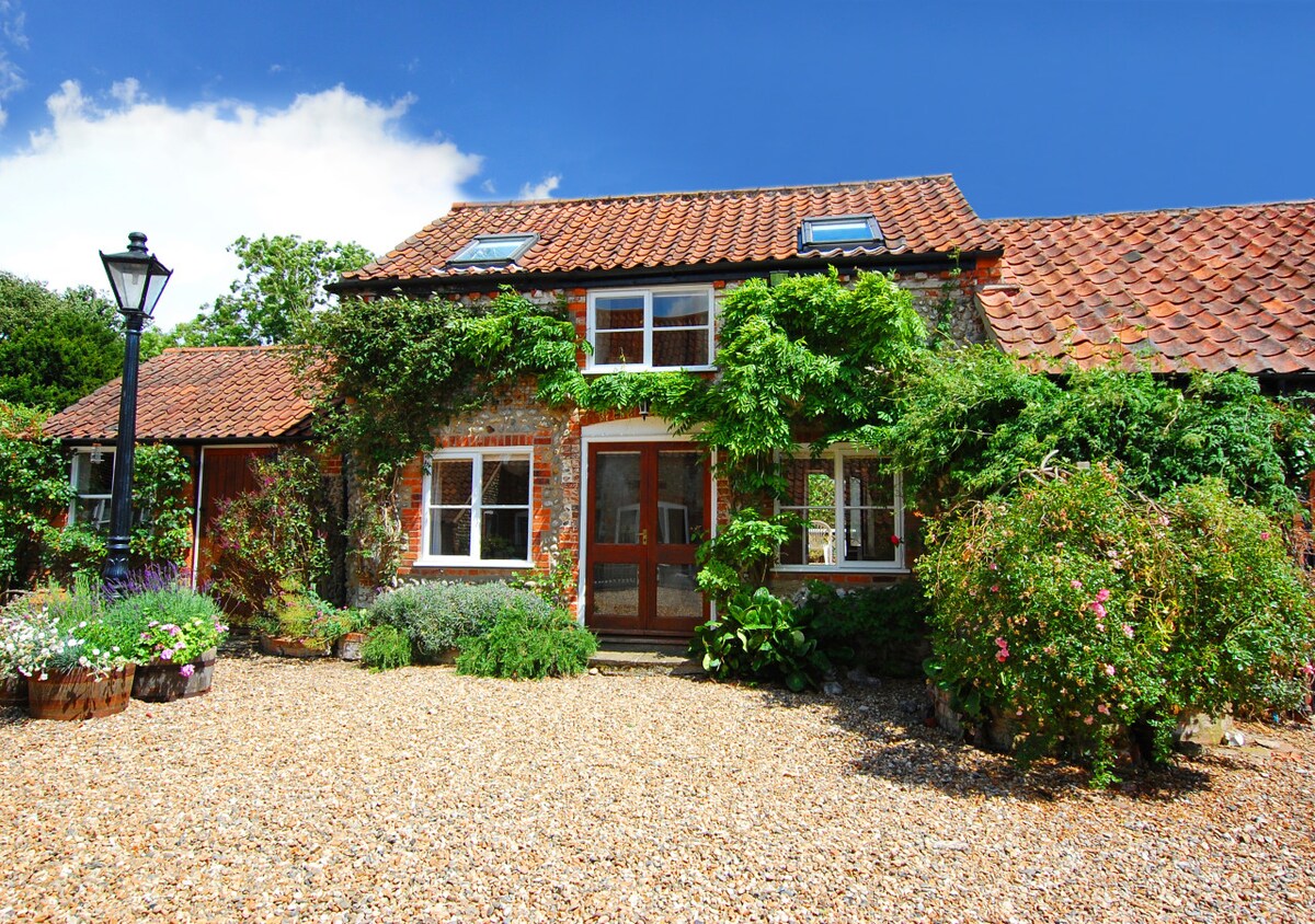 The charming cottage showcases a gravel pathway leading to its entrance, framed by lush greenery and flowering plants. The traditional brick facade is complemented by a red-tiled roof and multiple windows, allowing natural light to brighten the inviting facade.
