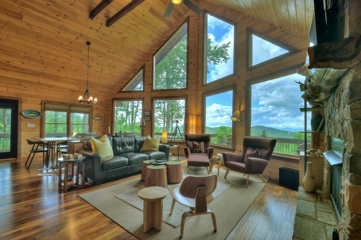 A well-appointed living area is highlighted by large triangular windows that offer expansive mountain views. The space includes a dark leather sofa, two modern chairs, and a wooden coffee table on a light area rug. A stone fireplace is situated to the right, enhancing the natural wood accents.