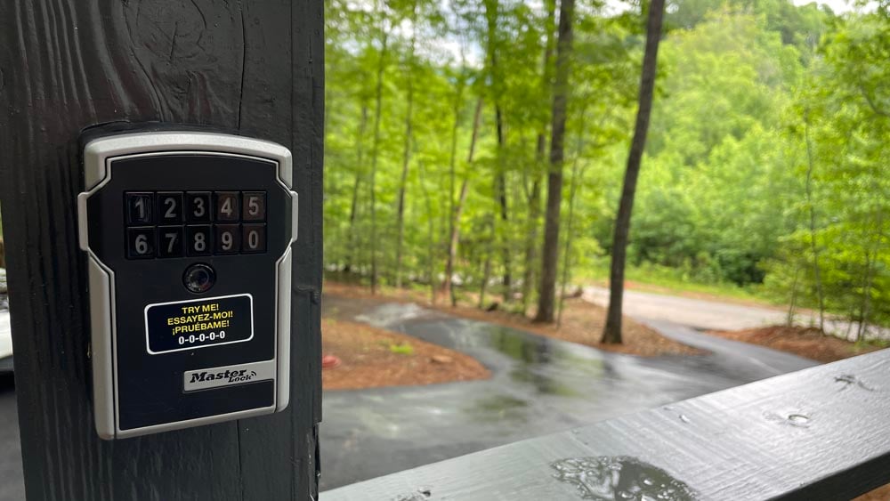 A keypad entry system is displayed on a wooden post. The lush green trees and winding driveway are visible in the background, creating a serene environment. The keypad features a simple number combination layout for secure access.